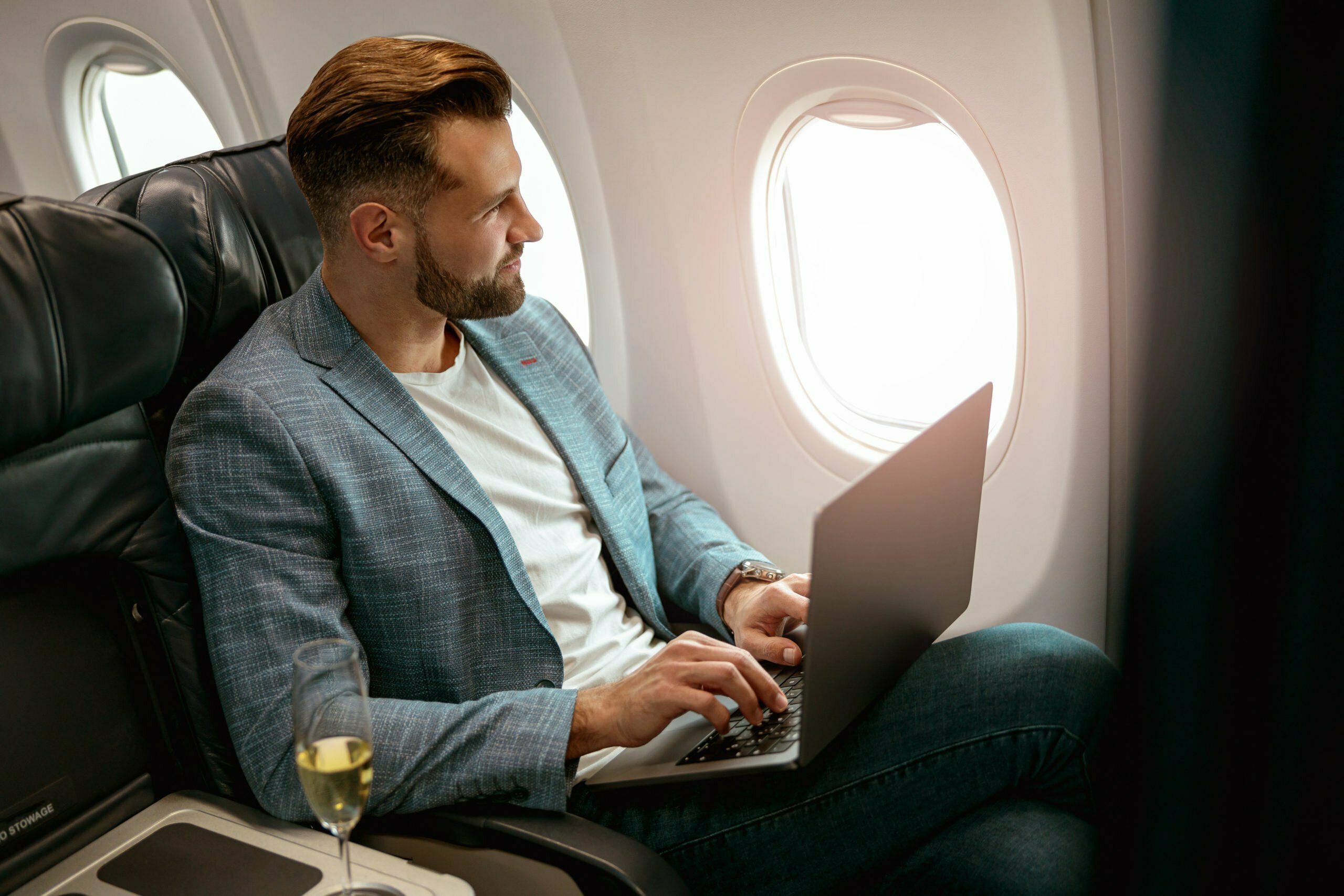 Bearded man sitting in passenger chair and looking out aircraft window while using modern notebook